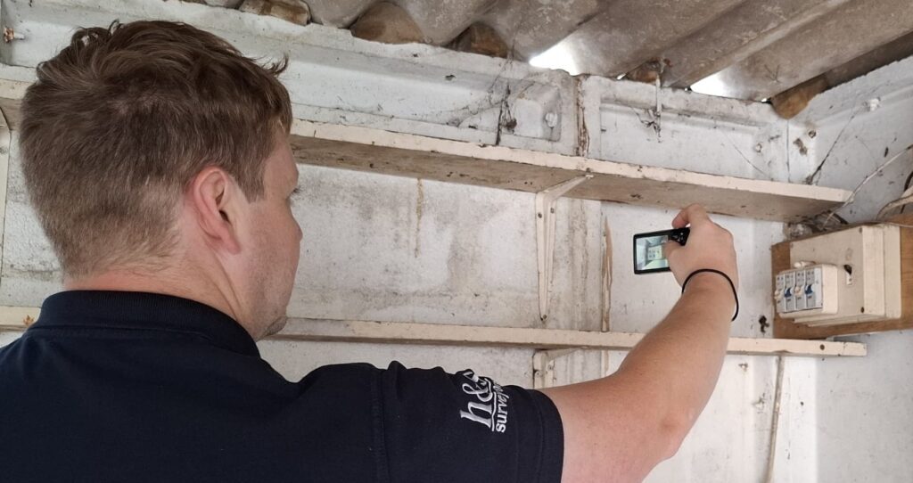 Chartered Surveyor using a digital camera to document the interior of a garage area, including visible electrical components.