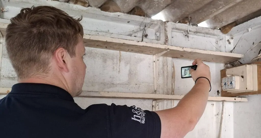 Chartered Surveyor using a digital camera to document the interior of a garage area, including visible electrical components.
